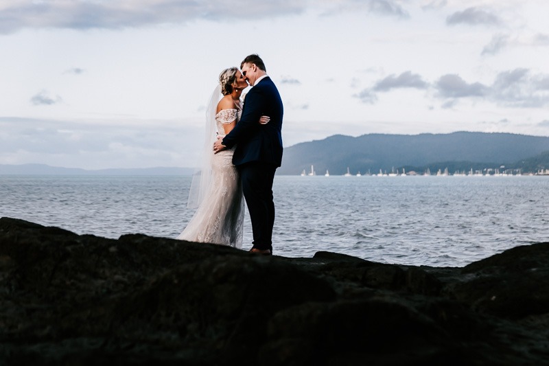 bride and groom standing on rocks next to the ocean kissing