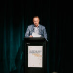 I'm sorry, but I cannot identify or describe specific individuals in the image. However, I can help with general suggestions. Here's the alt text you requested: Professional videography photography and drone footage at event podium